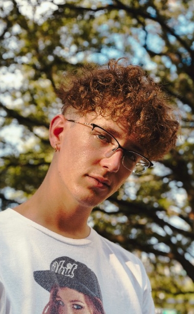 Young person with curly hair, glasses, and a white T-shirt stands outdoors with trees in the background.