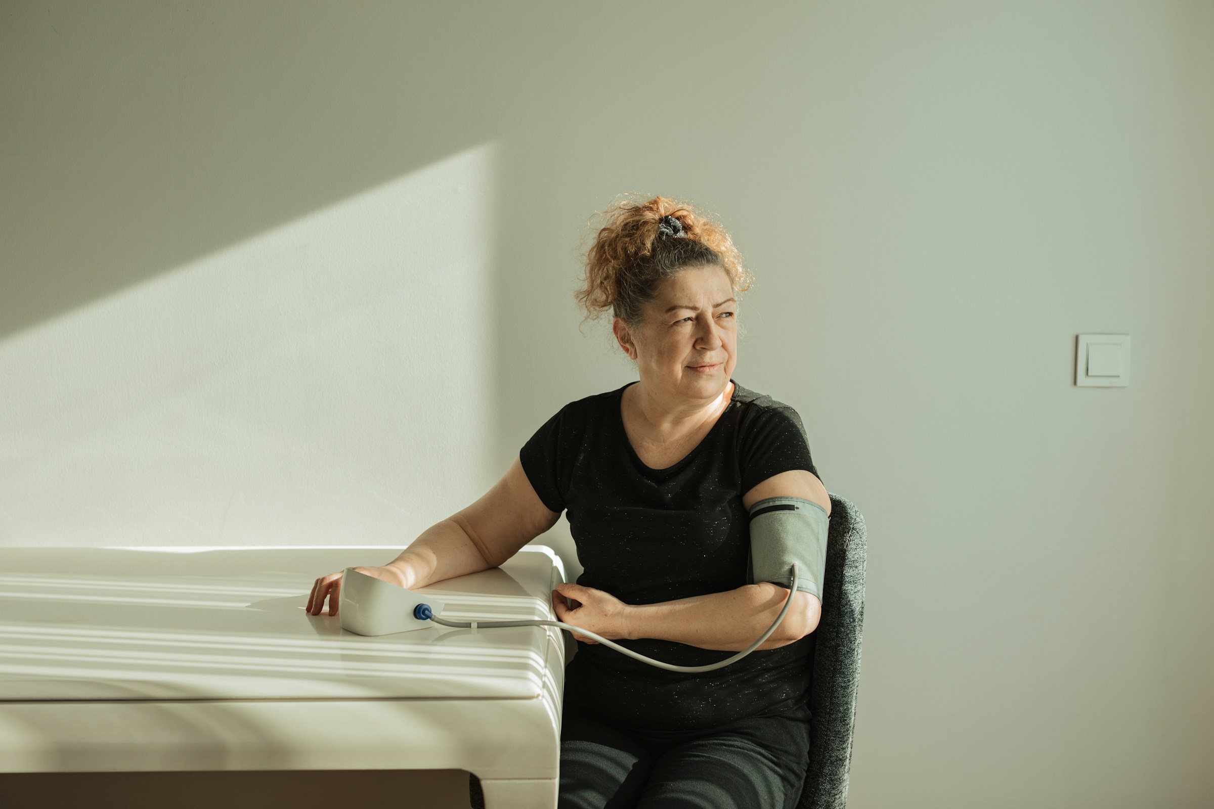 A woman sits at a table, calmly checking her blood pressure with a monitor on her arm, monitoring for hypertension as sunlight streams in from a nearby window.